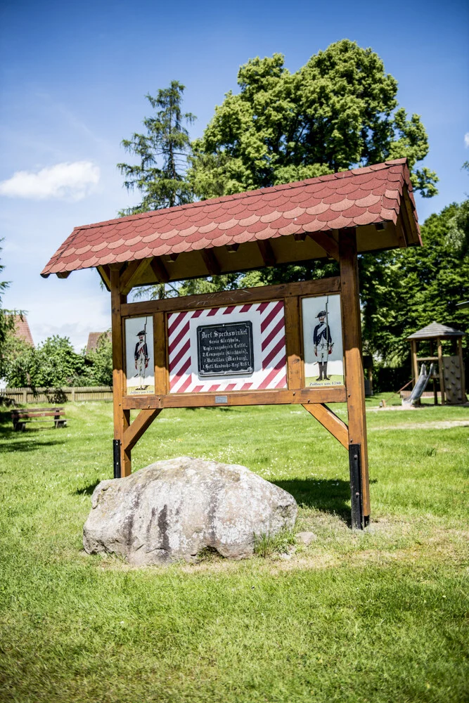 Impressionen aus Speckswinkel: überdachte Infotafel mit Findling auf grüner Wiese