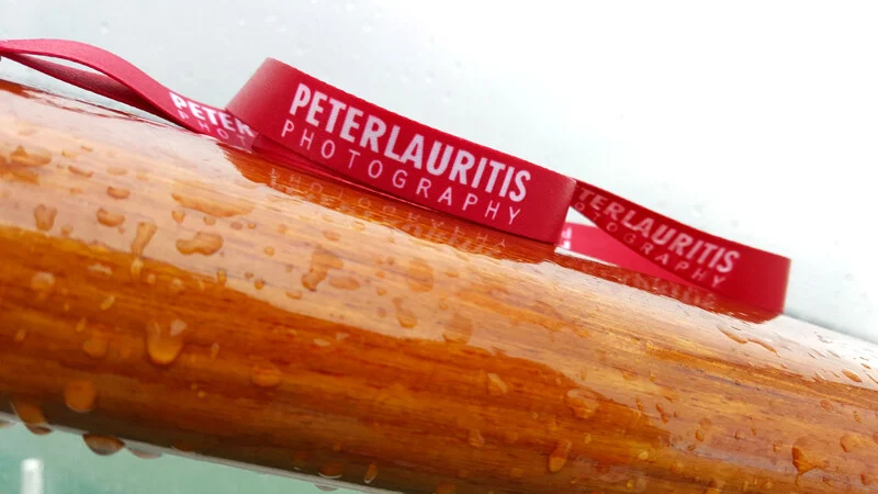 Schlüsselband mit Logo: zwei rote Bänder mit weißem Schriftzug auf nassem Holzgeländer