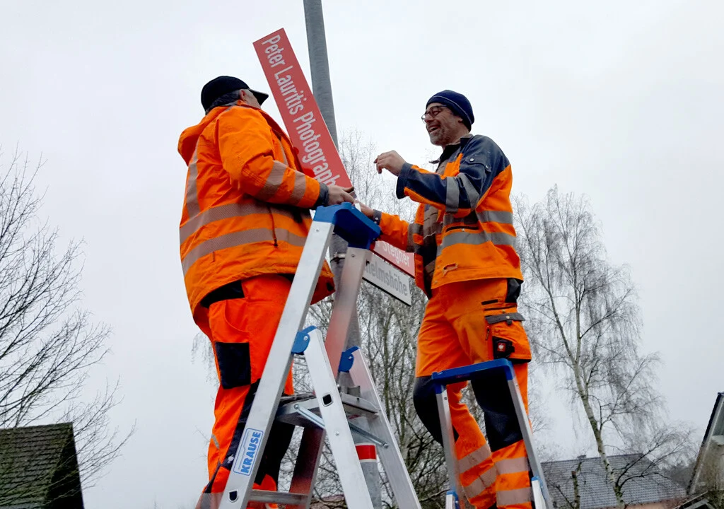 Neuer Wegweiser | Peter Lauritis Photography Wegweiser zum Fotostudio: zwei Helfer in Arbeitskleidung montieren das rote Schild auf Leitern