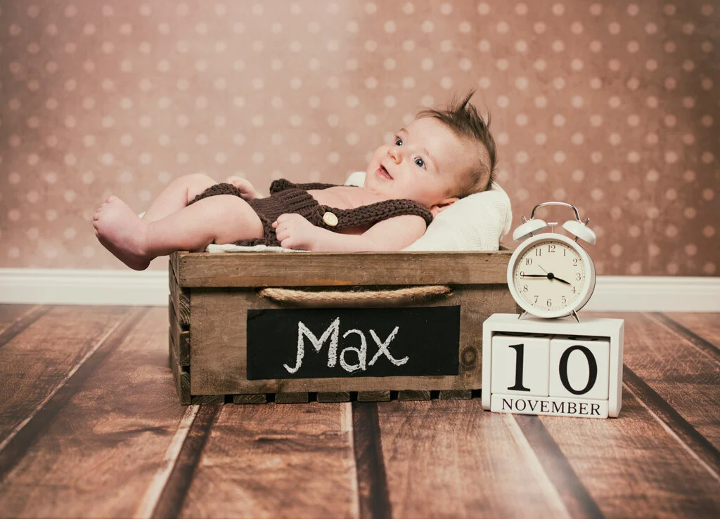 Baby in der Holzkiste | Peter Lauritis Photography Babyfotos mit Holzkiste: lächelndes Baby liegt in einer Holzkiste neben Wecker und Datumswürfeln