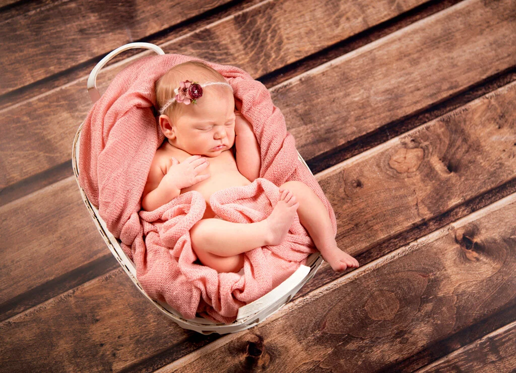 Neugeborenes im Körbchen | Peter Lauritis Photography Neugeborenenfotos im Körbchen: schlafendes Baby in rosa Tuch mit Blütenhaarband auf Holzboden