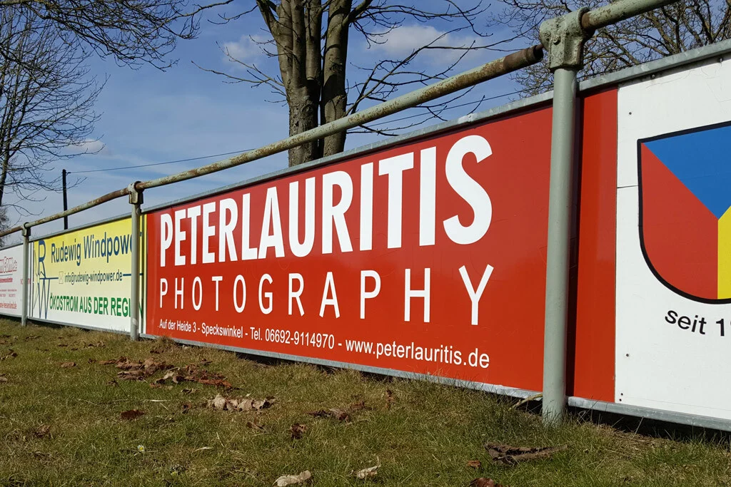 Bandenwerbung | Peter Lauritis Photography Bandenwerbung: rotes Banner von Peter Lauritis Photography am Geländer eines Sportplatzes