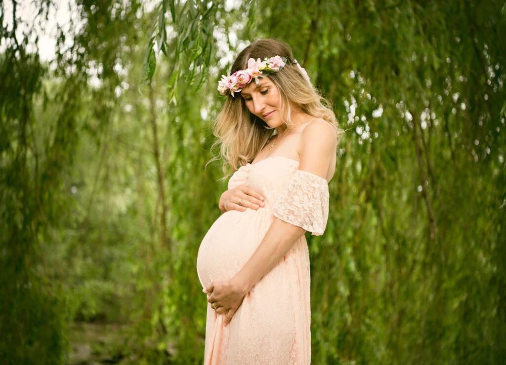 Babybauch und Blumenkranz: Schwangere im rosa Spitzenkleid mit Blütenkranz vor einer Trauerweide