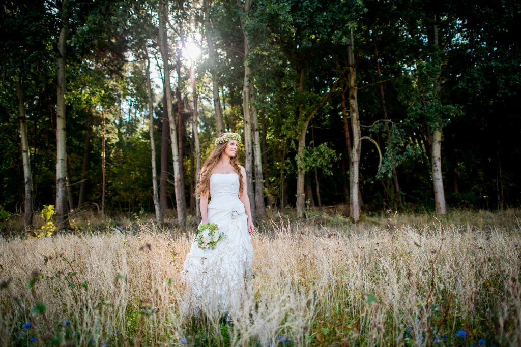 Frau im Brautkleid mit Strauß steht auf einer Wiese vor sonnigem Wald