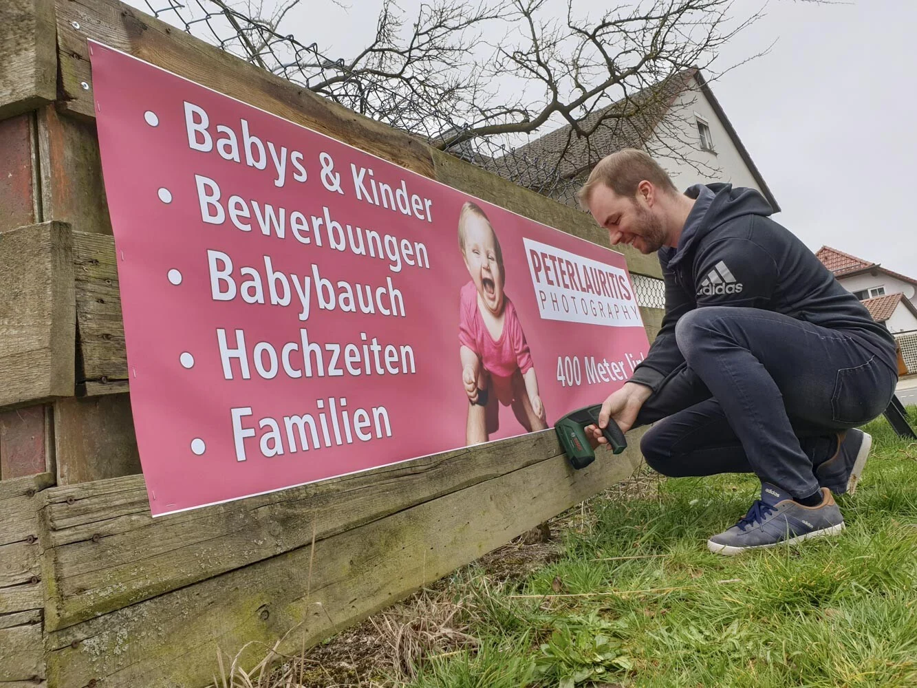 Mann schraubt ein pinkes Werbebanner mit dem Akkuschrauber an einen Holzzaun
