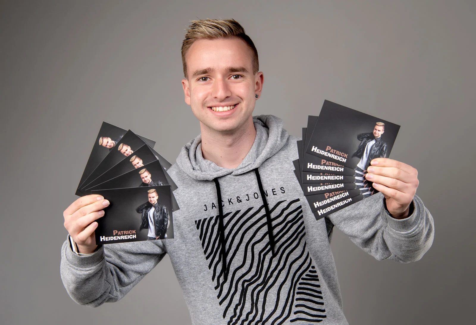Lachender junger Mann im grauen Hoodie hält zwei Stapel seiner Autogrammkarten