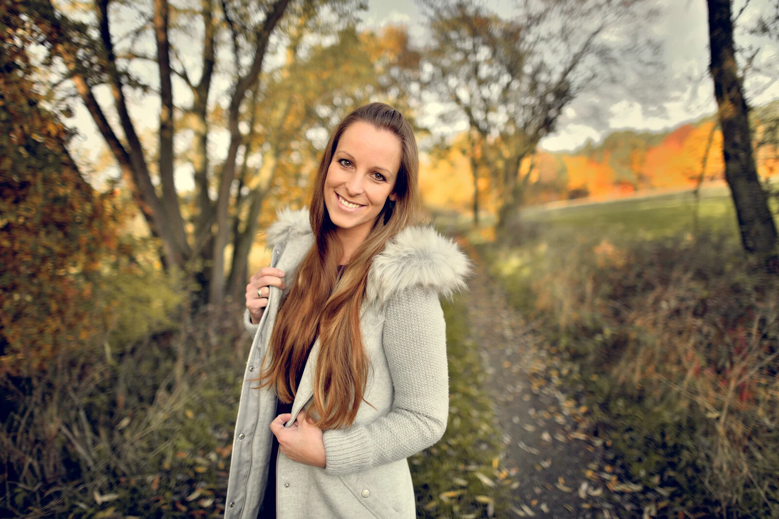Lächelnde Frau in cremefarbener Kapuzenjacke steht an einem herbstlichen Feldweg