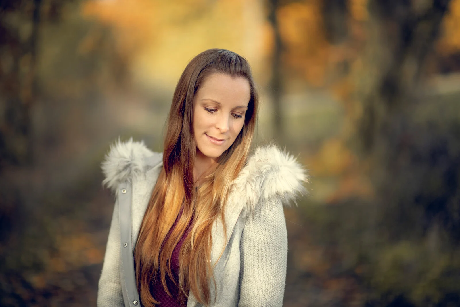 Herbstportraits: junge Frau mit langen Haaren und heller Jacke blickt vor goldenem Laub nach unten