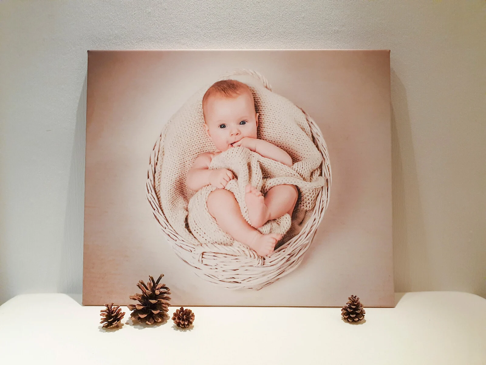 Leinwände in Handarbeit: Babyfoto im Weidenkorb auf Leinwand, davor liegen Tannenzapfen