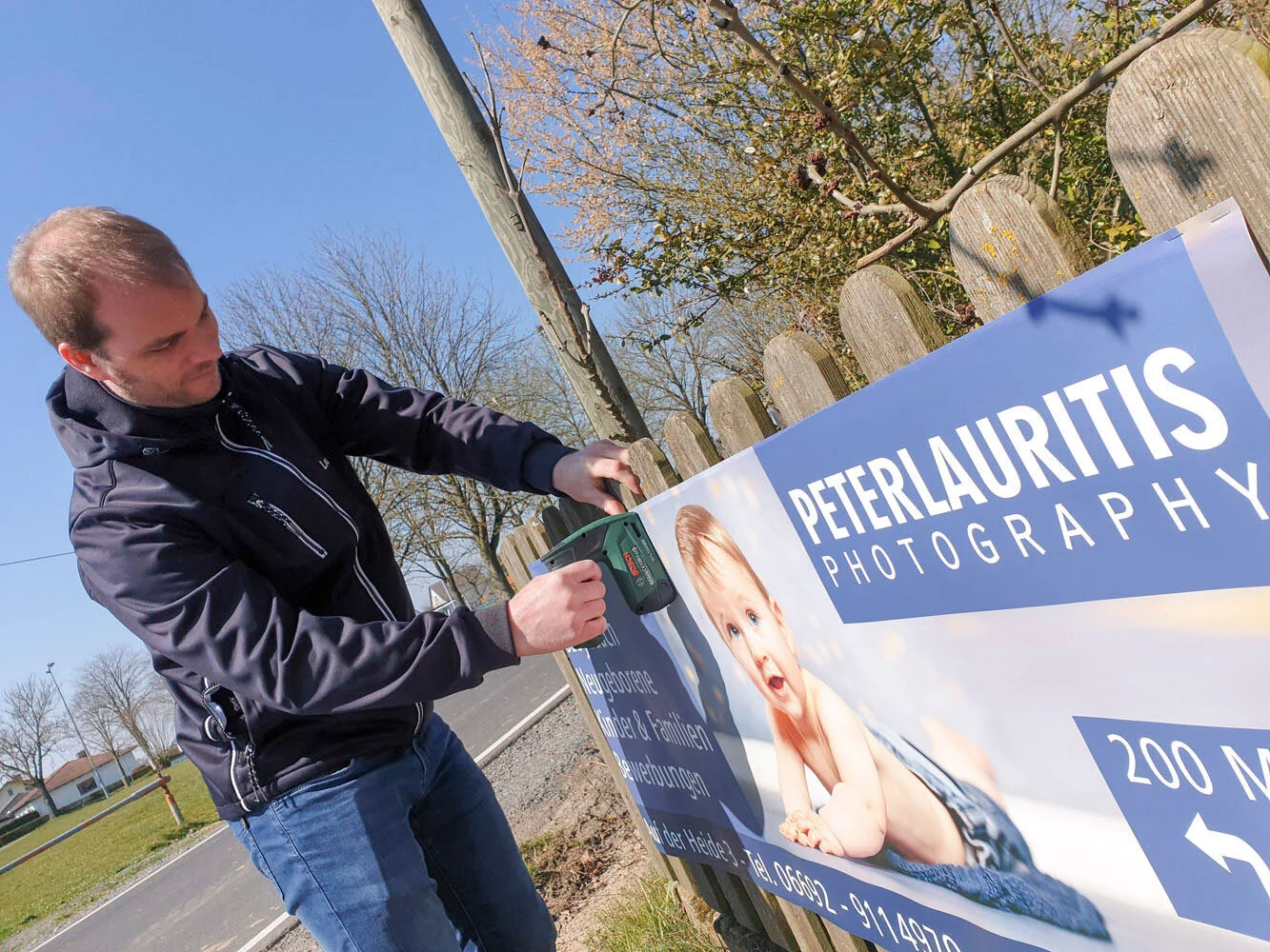 Werbebanner mit Babyfoto wird mit einem Akkuschrauber an einem Holzzaun befestigt