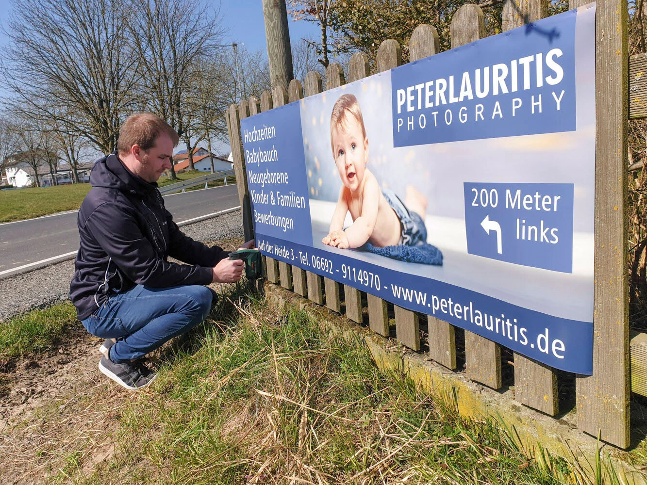 Mann kniet neben einem großen Werbebanner von Peter Lauritis Photography an einem Holzzaun