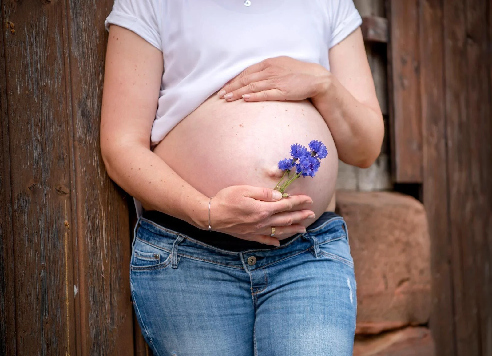 Schwangere hält Kornblumen an ihren Babybauch, im Hintergrund altes Holz