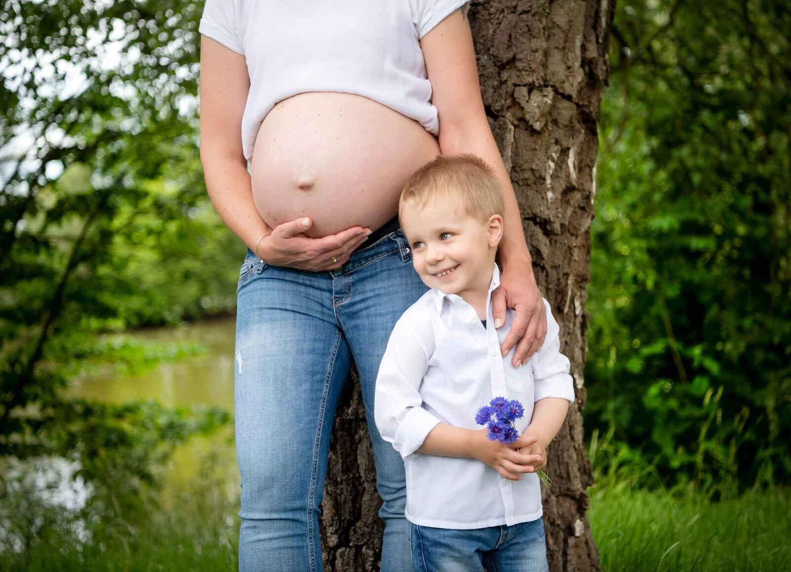 Babybauchfotoshooting: kleiner Junge mit Kornblumen lehnt lächelnd an Mamas Bauch