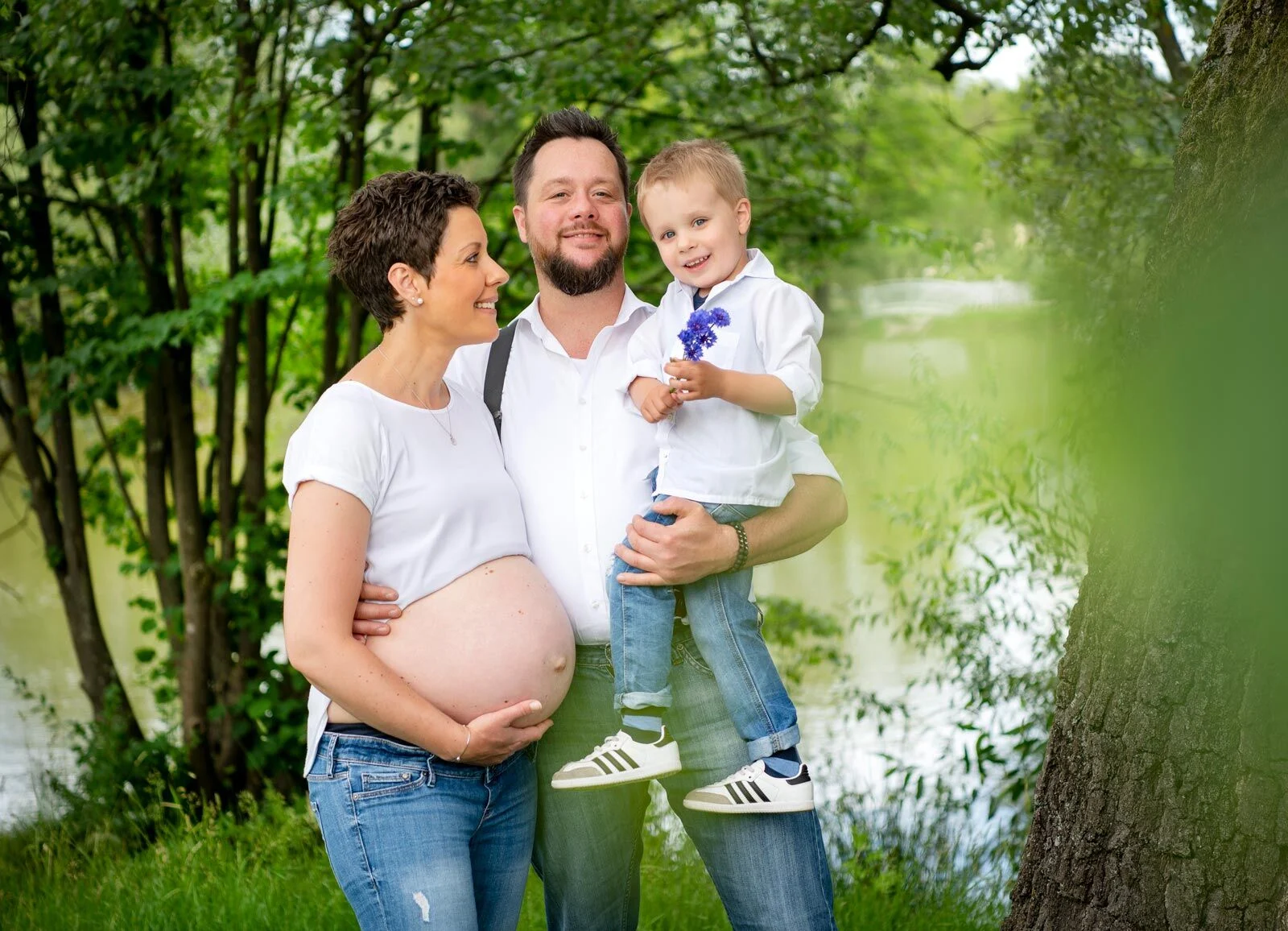 Babybauchfotoshooting am Wasser: Paar in Weiß, der Vater trägt den kleinen Sohn