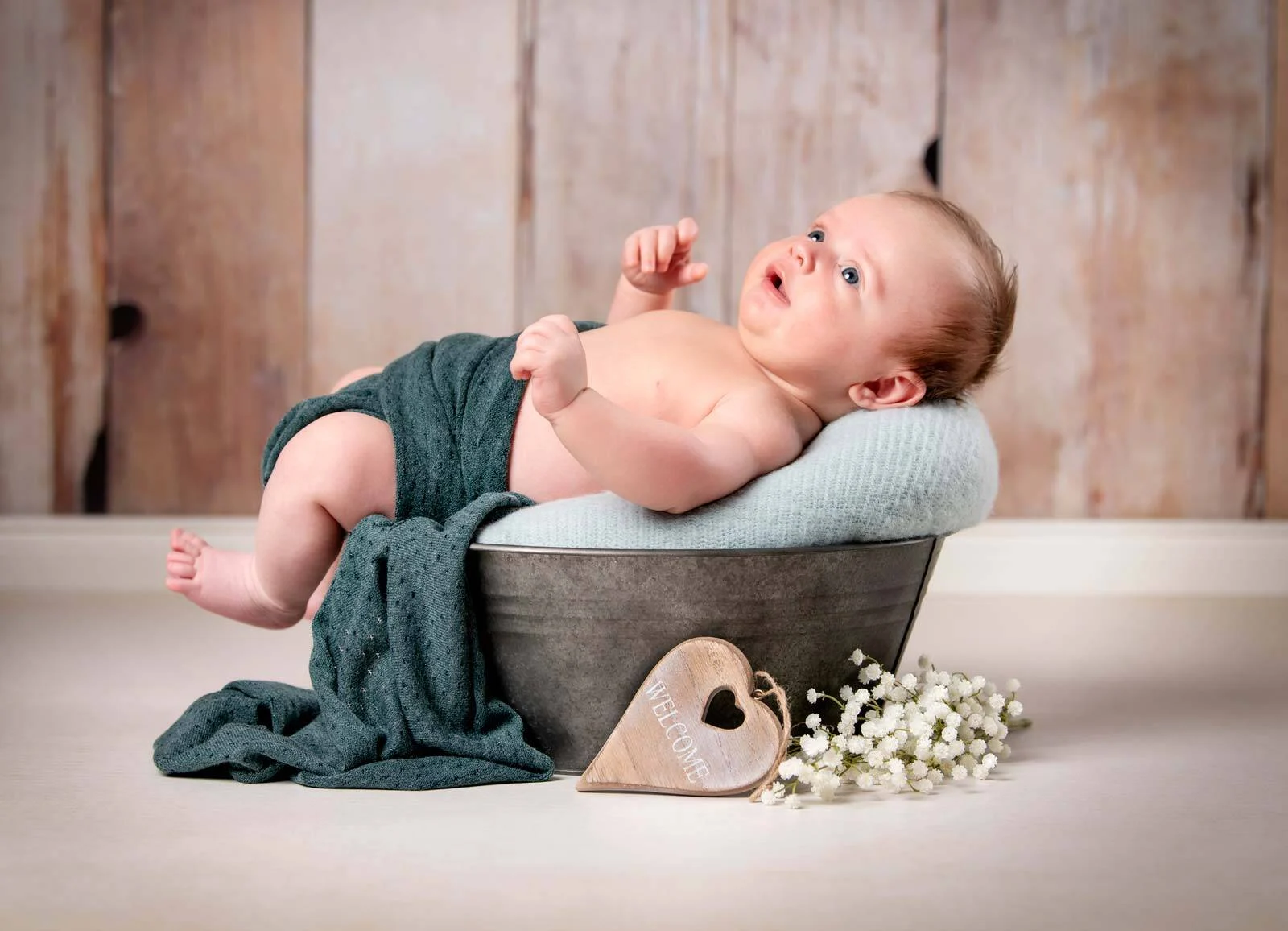 Baby liegt mit grüner Decke in einer Zinkwanne und schaut nach oben, daneben ein Holzherz
