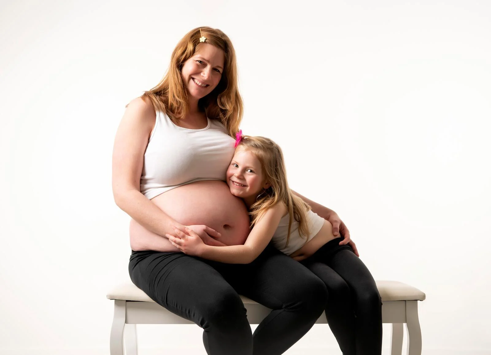 Kugelzeit: Mädchen kuschelt sich an den Babybauch der Mutter auf einer weißen Bank