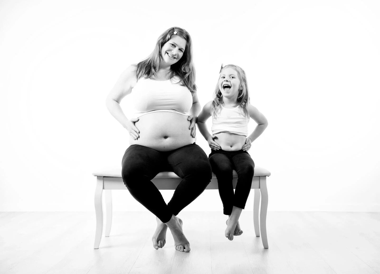 Schwarz-Weiß-Foto: Schwangere und ihre Tochter sitzen lachend auf einer Bank und halten den Bauch