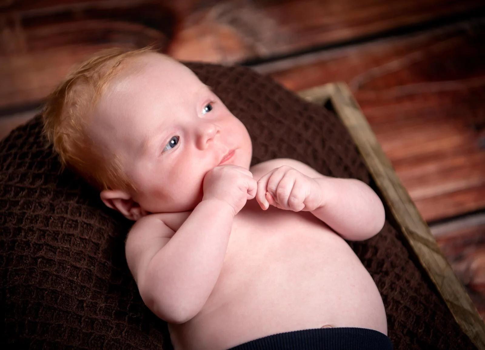 Baby auf brauner Strickdecke hält die Händchen ans Kinn und schaut zur Seite