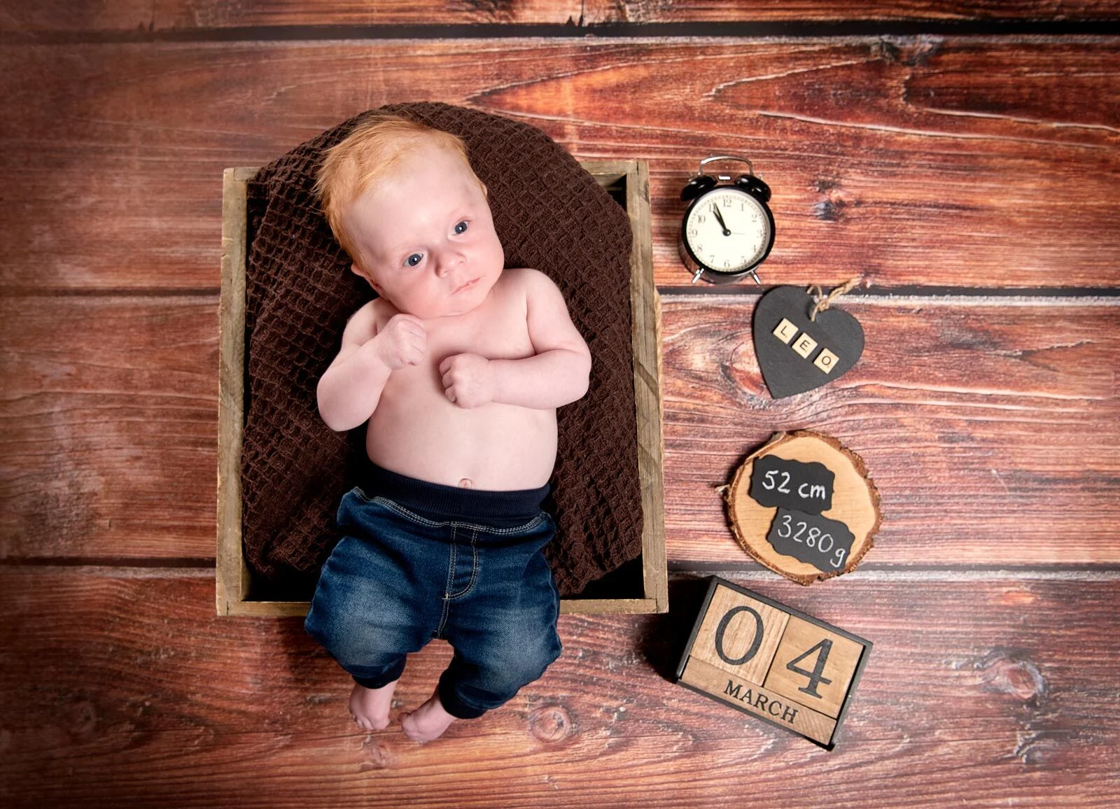 Baby in Jeans liegt in einer Holzkiste, daneben Wecker, Holzherz, Holzscheibe und Datumswürfel