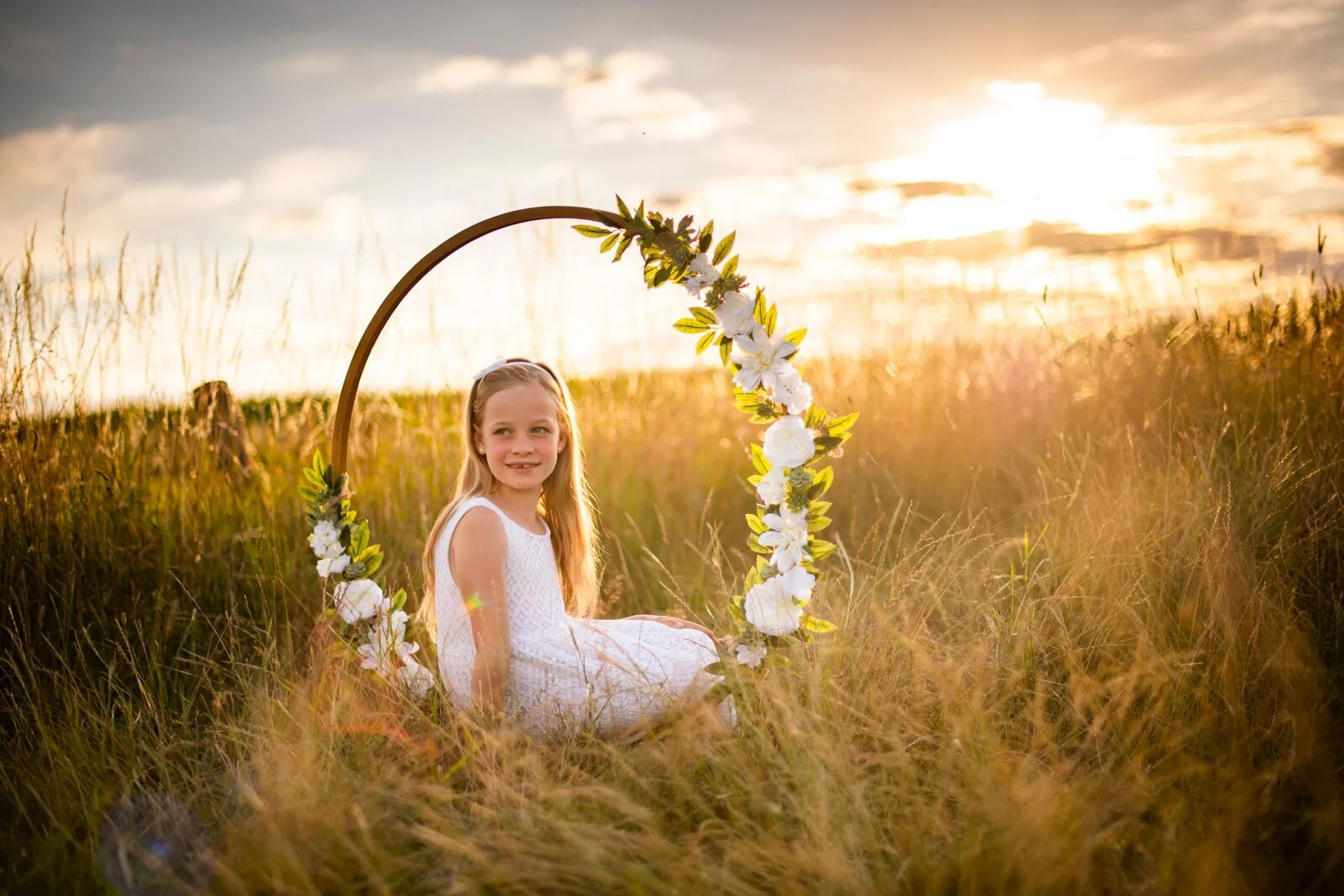Mädchen im weißen Kleid sitzt in einem Blumenring im Gras vor der untergehenden Sonne