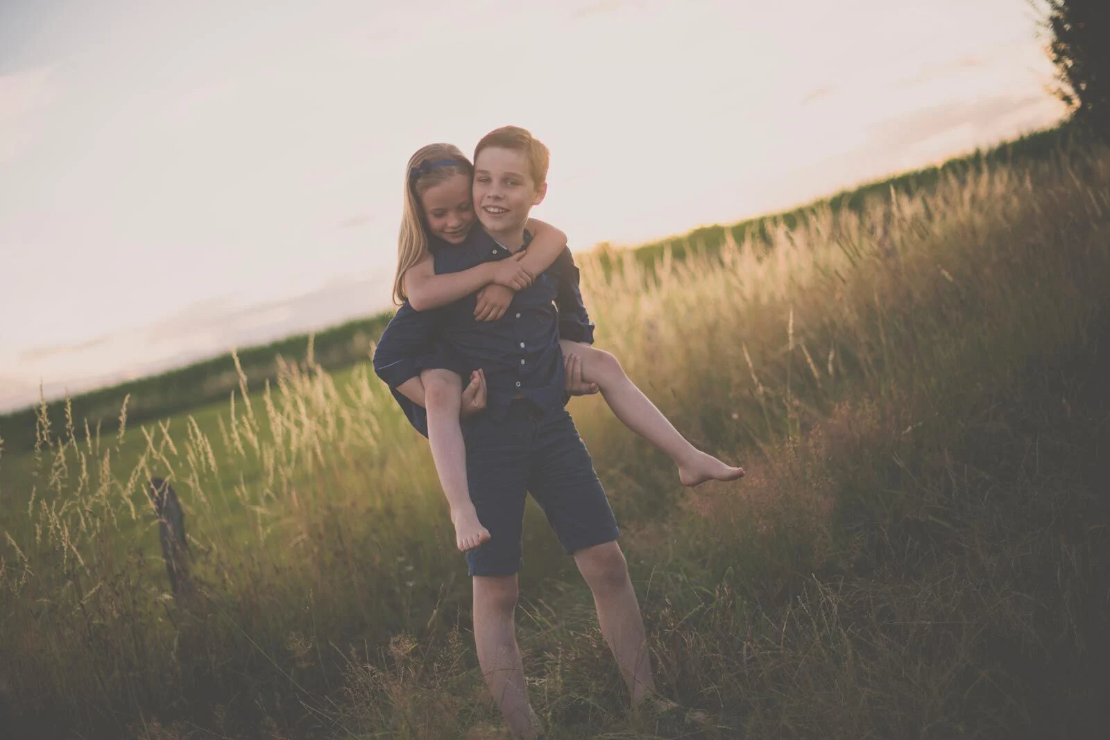 Junge trägt seine kleine Schwester huckepack über eine Wiese am Hang