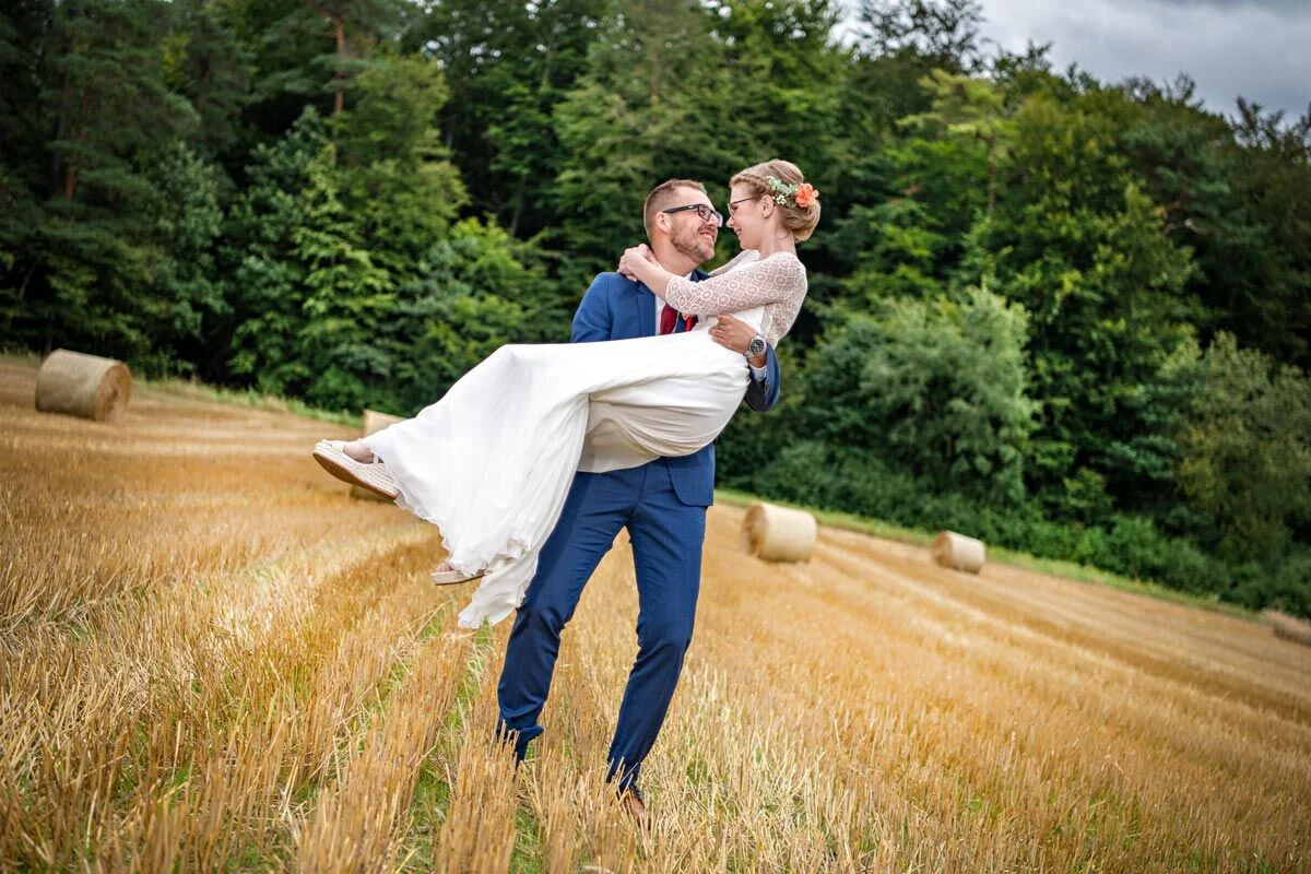 Traumhochzeit: Bräutigam trägt die Braut auf Händen über ein Stoppelfeld mit Strohballen