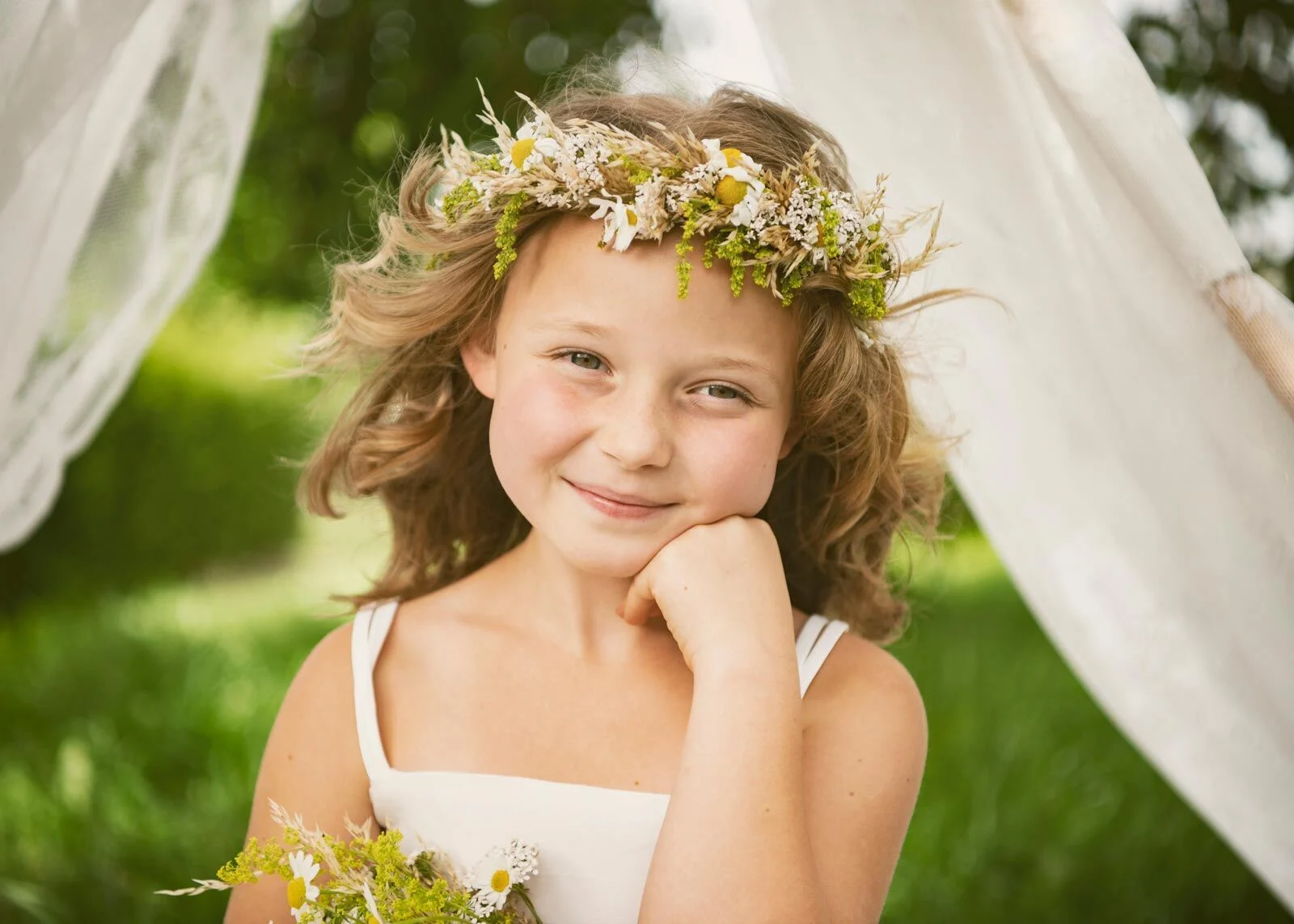 Mädchen mit lockigem Haar und Blumenkranz stützt das Kinn auf die Hand