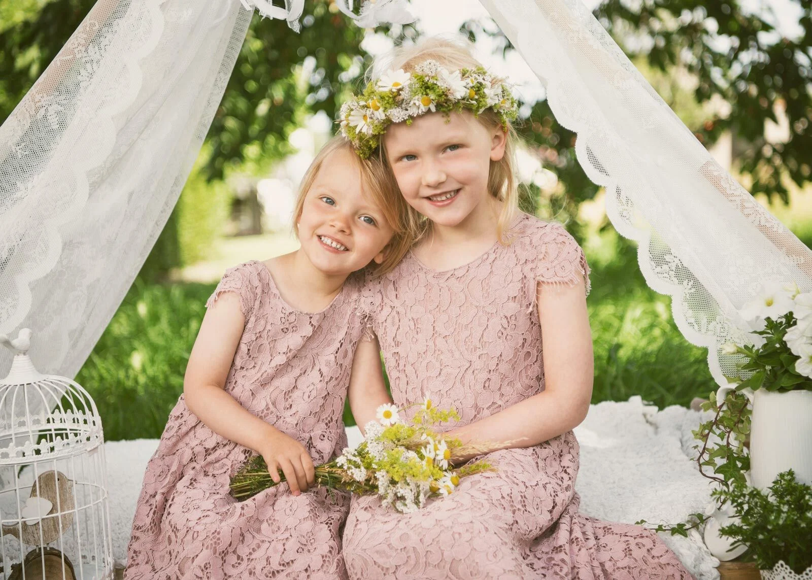 Zwei Mädchen in rosa Spitzenkleidern mit Blumenkranz sitzen vor weißen Vorhängen