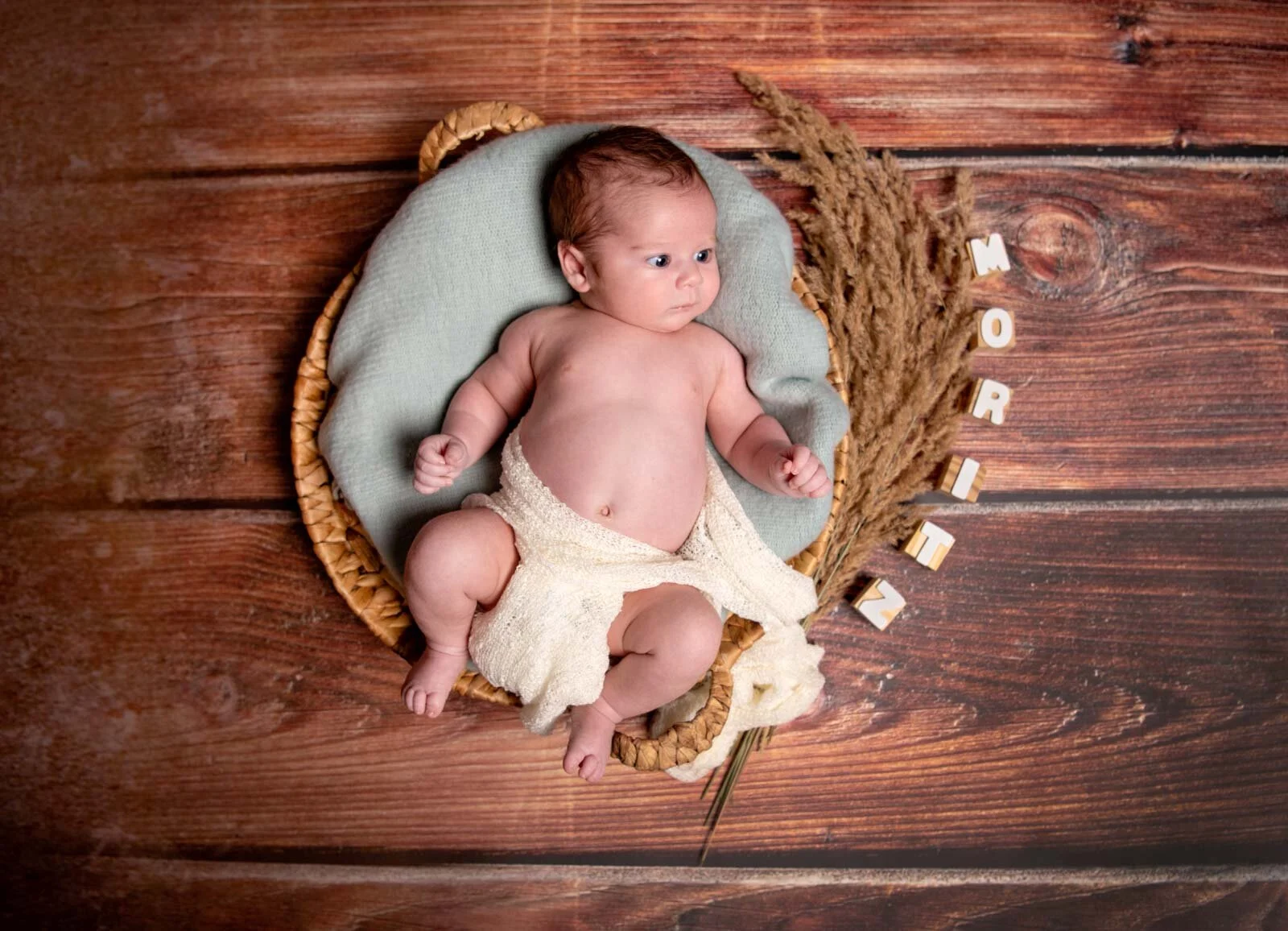 Babyshooting von oben: Baby liegt im runden Körbchen auf Holzboden neben Gräsern