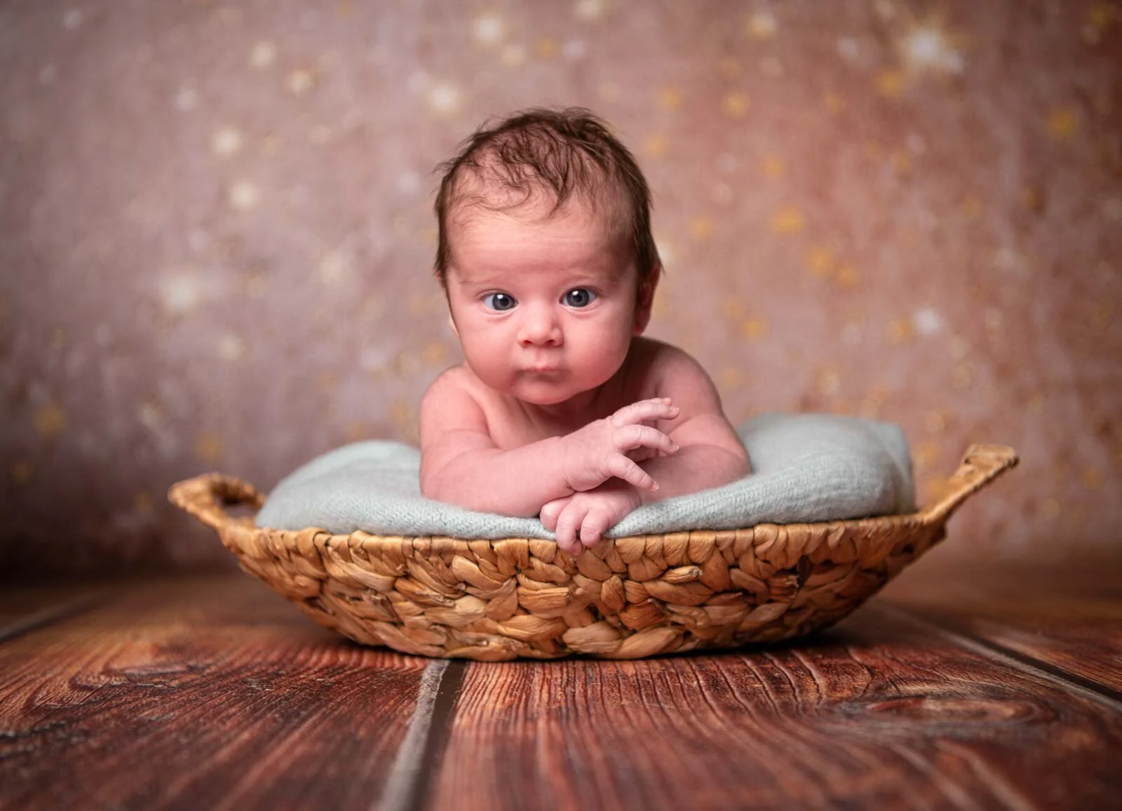 Baby liegt auf dem Bauch in einem flachen Korb und schaut mit großen Augen in die Kamera