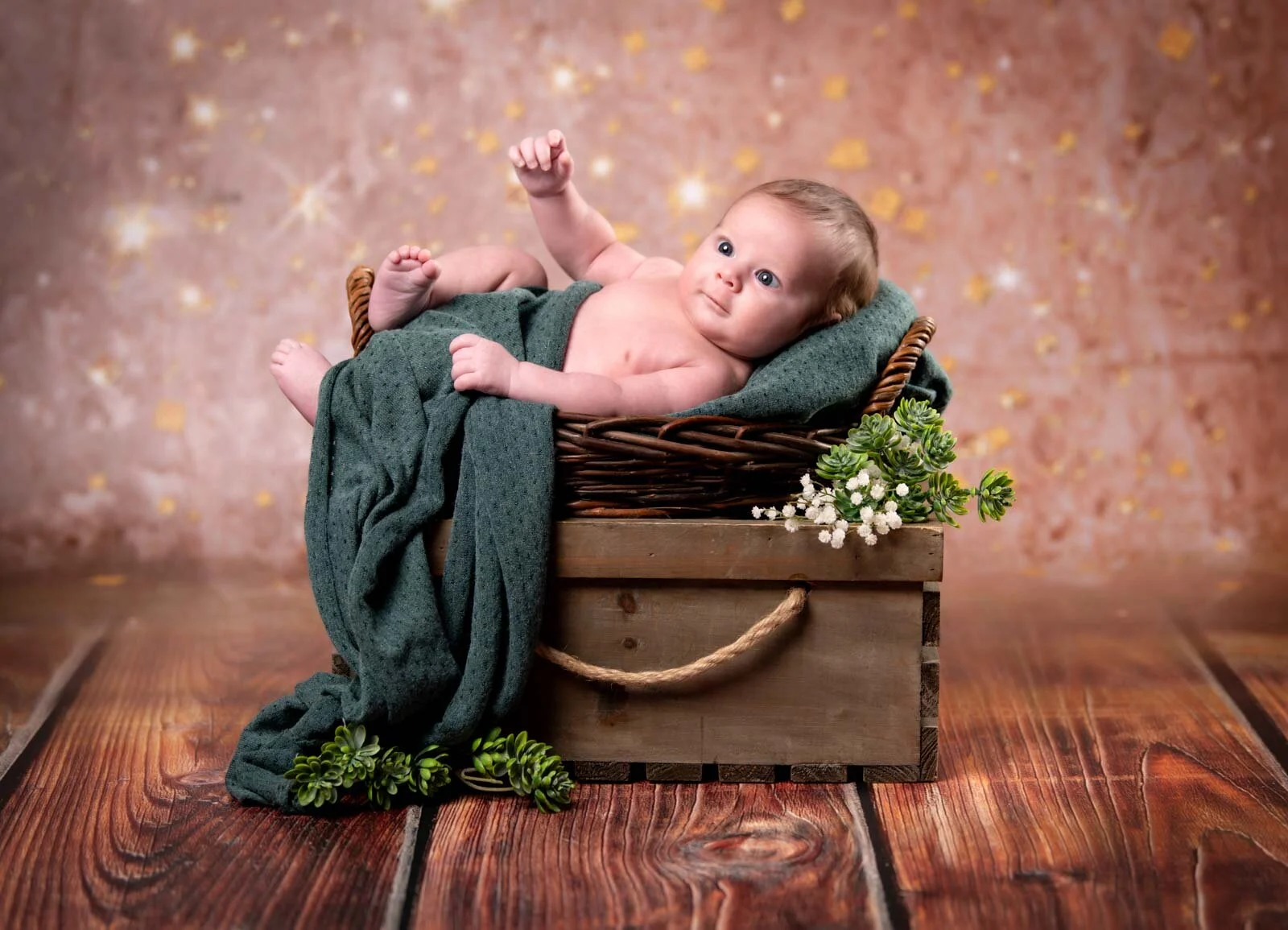 Baby liegt in einem Korb auf einer Holzkiste und streckt ein Ärmchen hoch