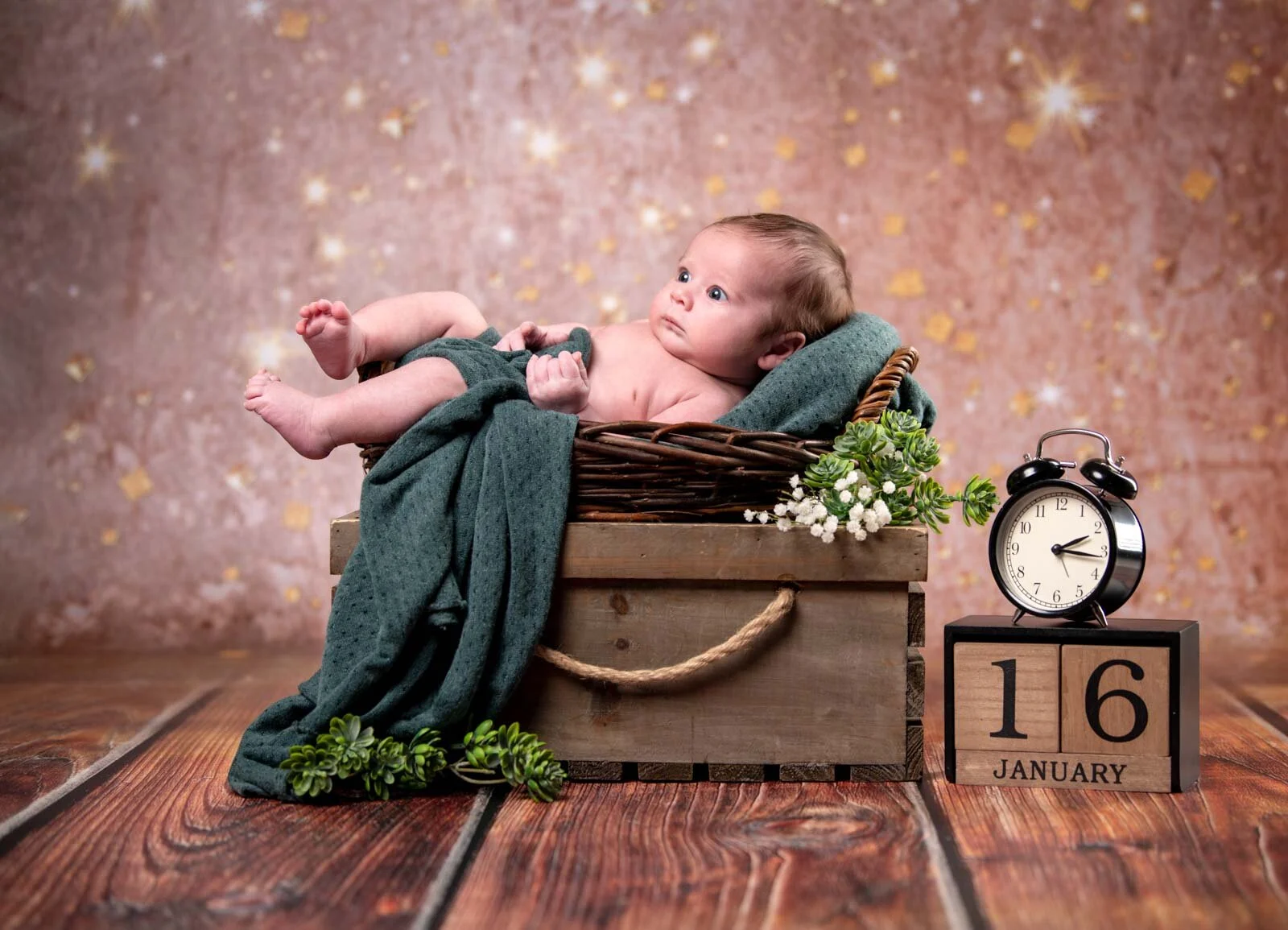 Baby in einer Holzkiste mit grüner Decke, daneben Wecker und Holzwürfel