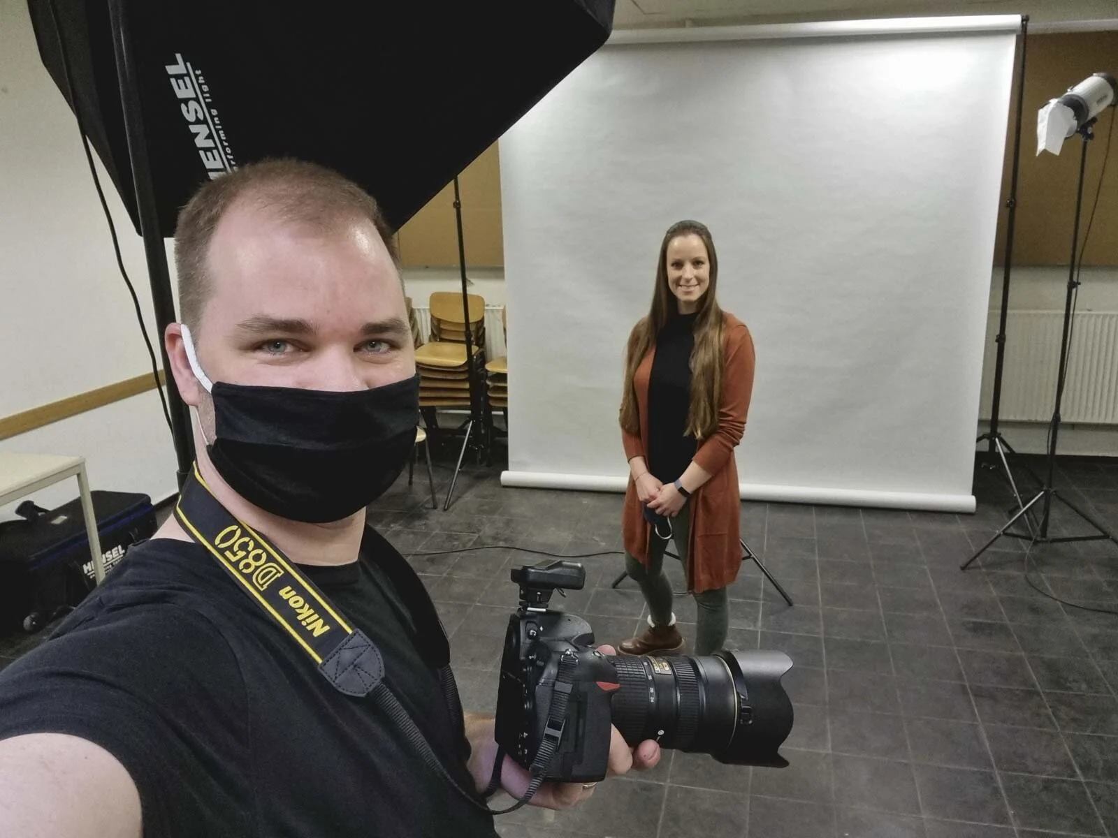 Fotograf mit Maske macht ein Selfie, dahinter steht eine Frau vor dem mobilen Studiohintergrund