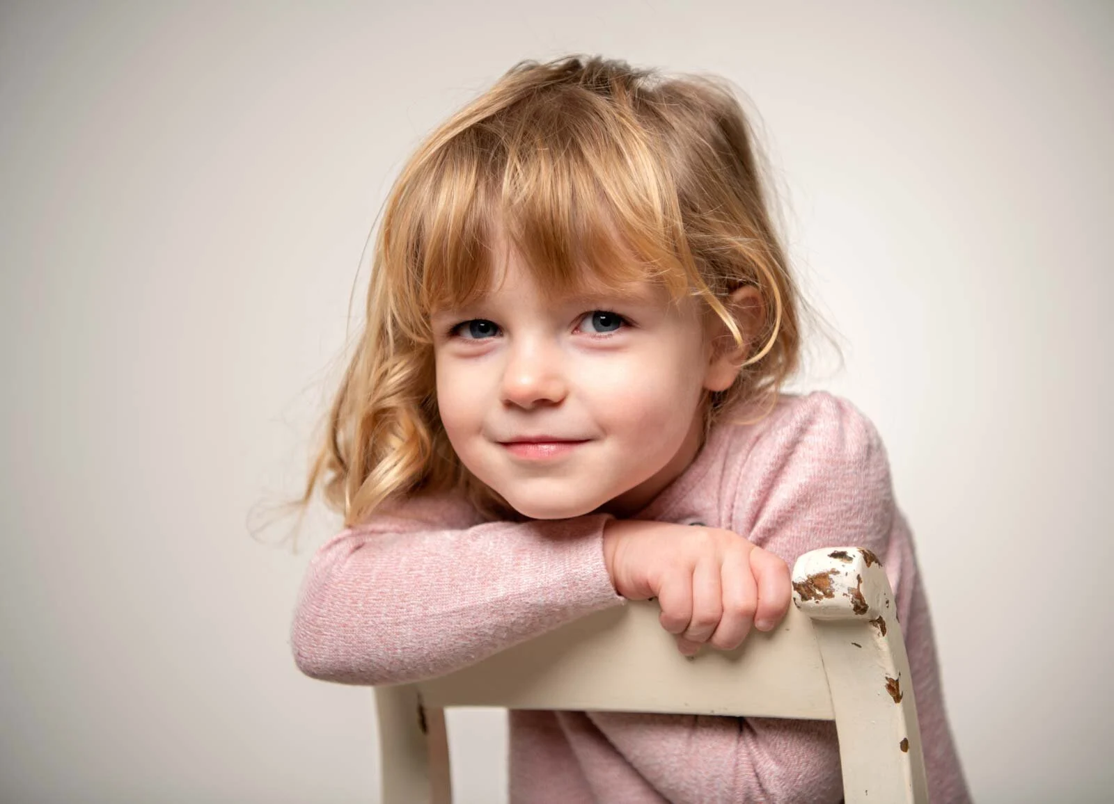 Kleines blondes Mädchen lehnt sich lächelnd über die Lehne eines weißen Stuhls