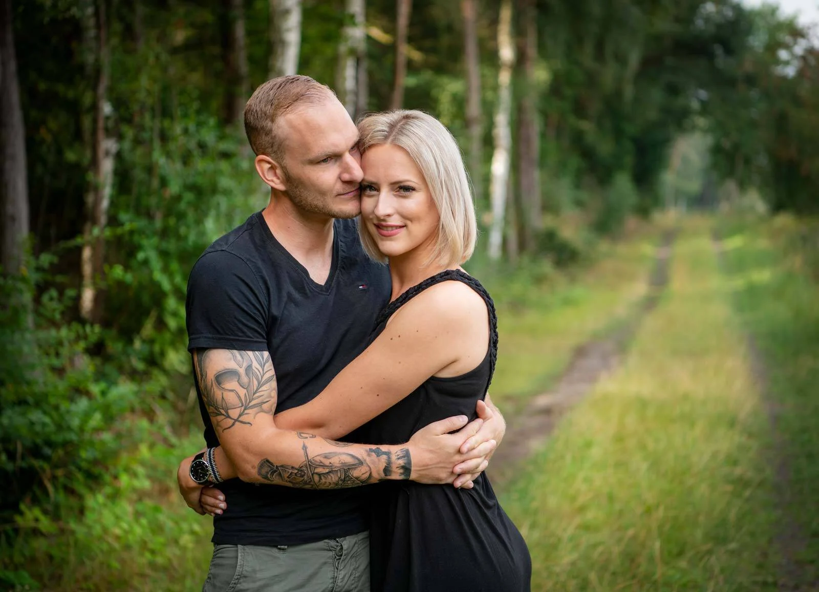 Paar umarmt sich auf einem grünen Waldweg und lächelt in die Kamera
