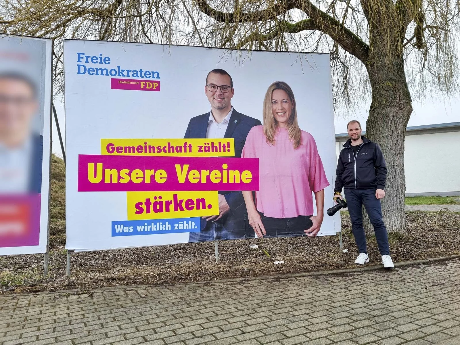 Fotograf steht neben einem Plakat mit einem Mann und einer Frau unter einer Weide