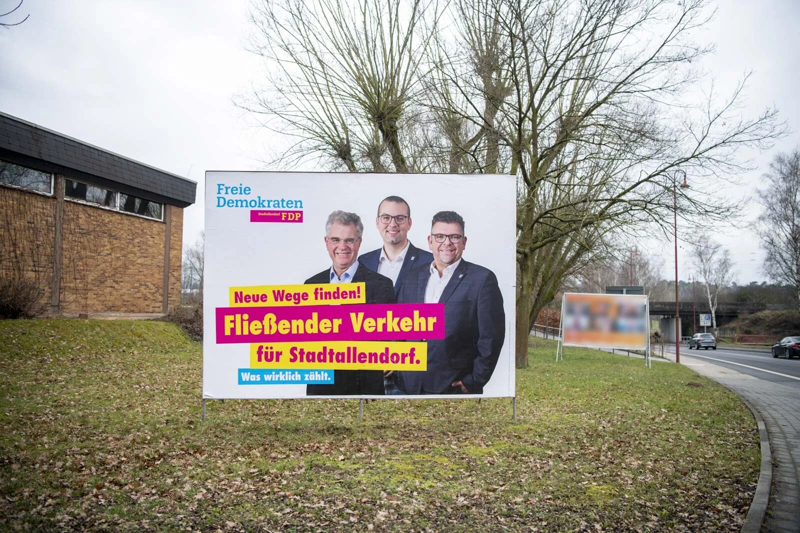 Großes Plakat mit drei Kandidaten am Straßenrand zwischen kahlen Bäumen