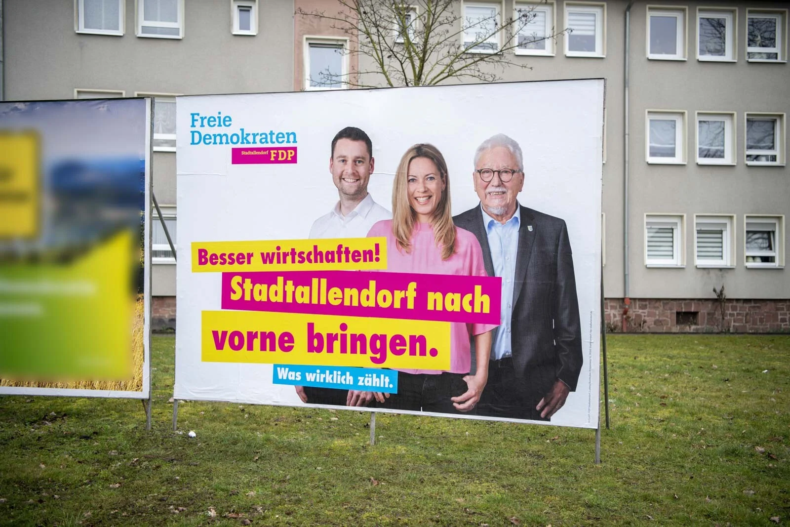 Wahlplakat mit zwei Männern und einer Frau vor einem Wohnhaus auf einer Wiese