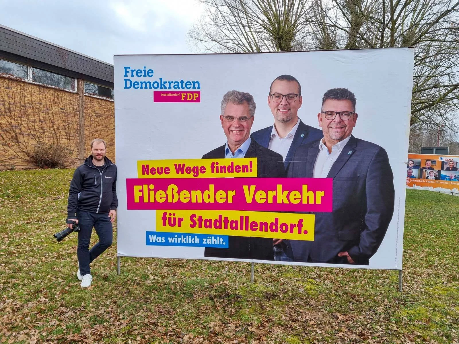 Fotograf mit Kamera steht neben einem großen Wahlplakat mit drei Kandidaten