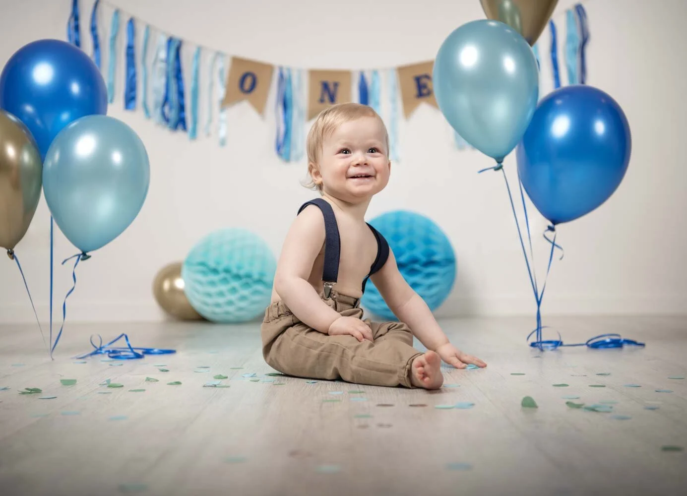Cake Smash Fotoshooting: lachender Junge mit Hosenträgern zwischen blauen Luftballons