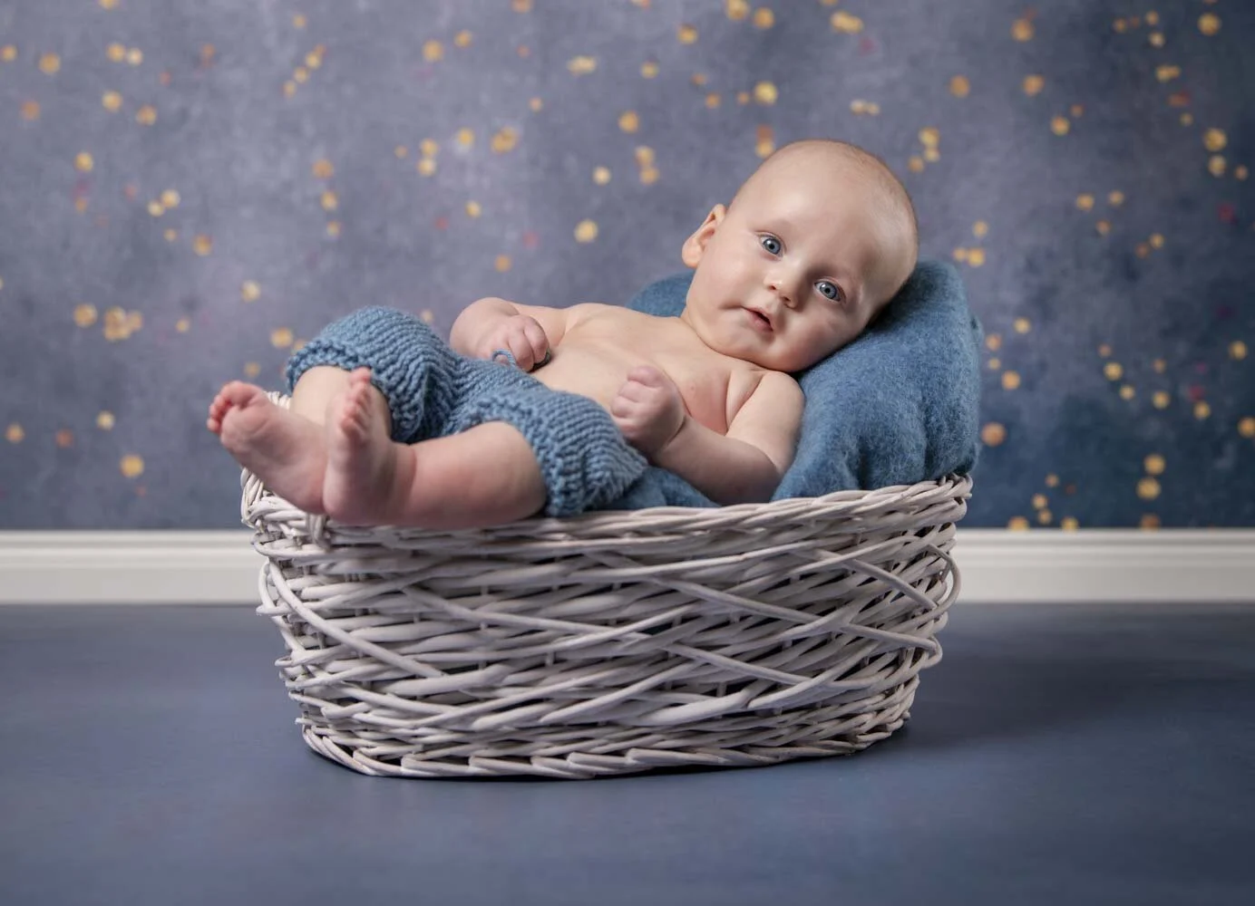 Baby in blauer Strickhose liegt entspannt in einem weißen Weidenkorb