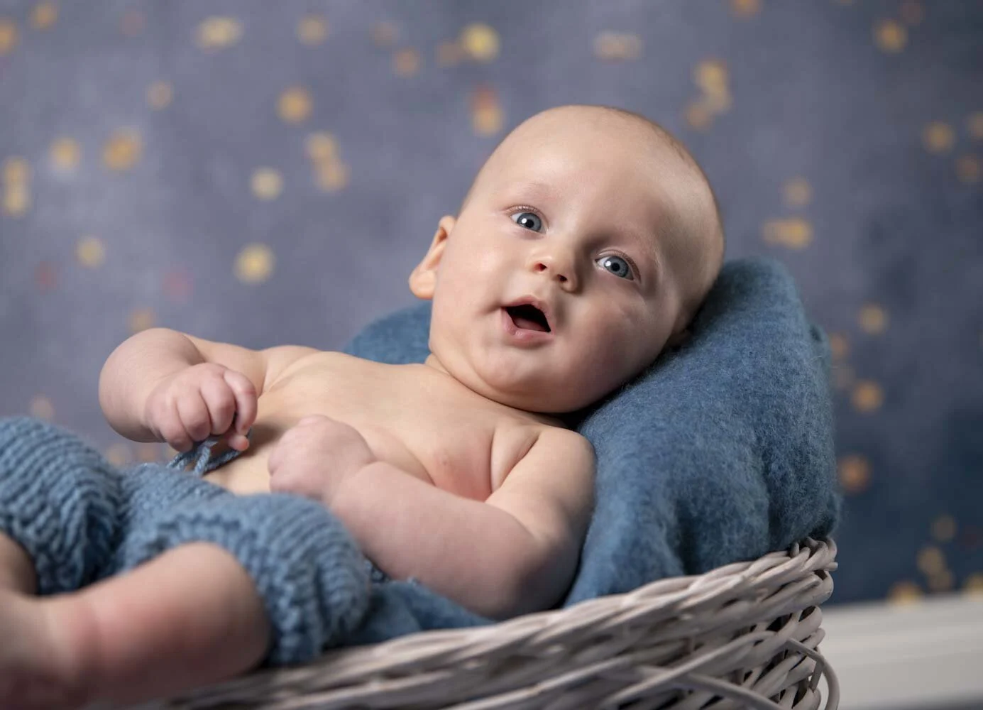 Baby liegt staunend in einem Korb auf blauer Decke vor goldenen Lichtpunkten