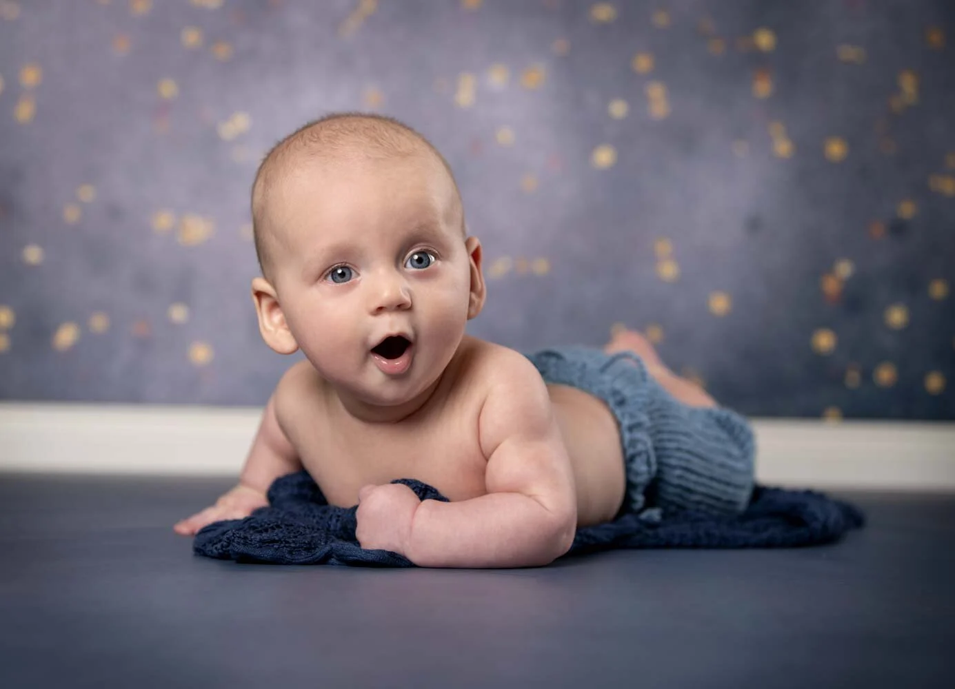 Baby liegt auf dem Bauch auf einer blauen Decke und schaut mit offenem Mund