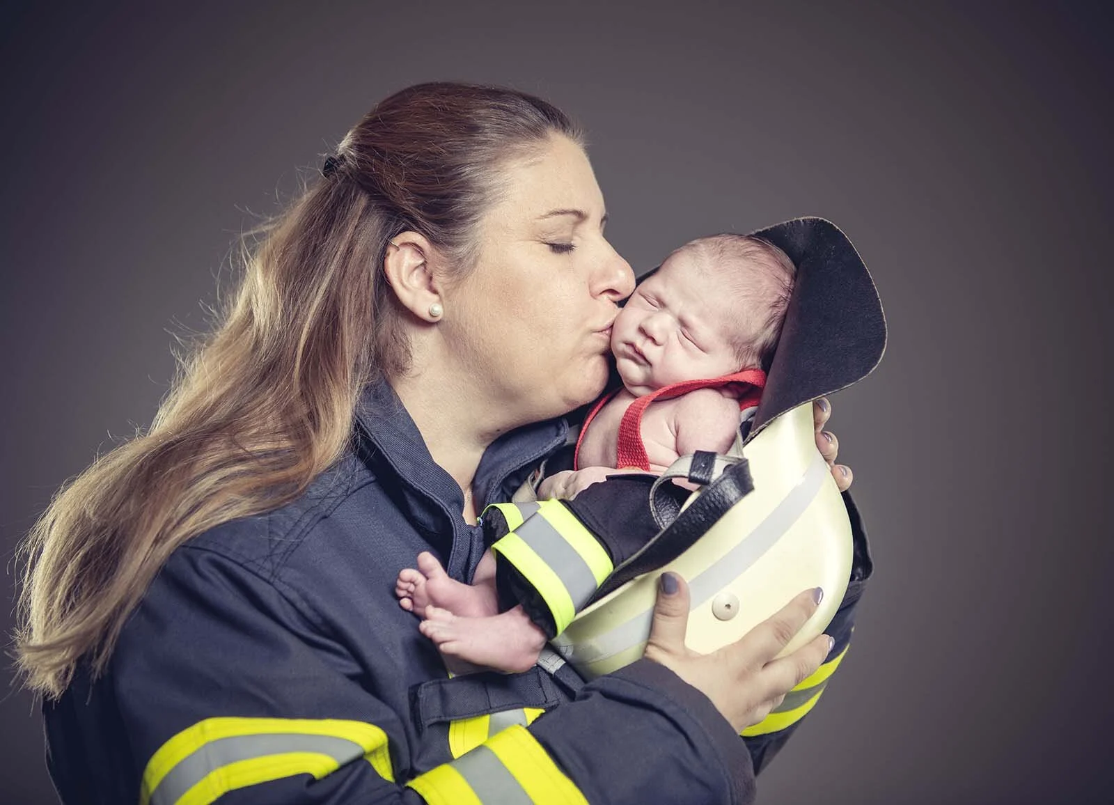 Mutter in Einsatzjacke küsst ihr schlafendes Neugeborenes, das in einem Feuerwehrhelm liegt