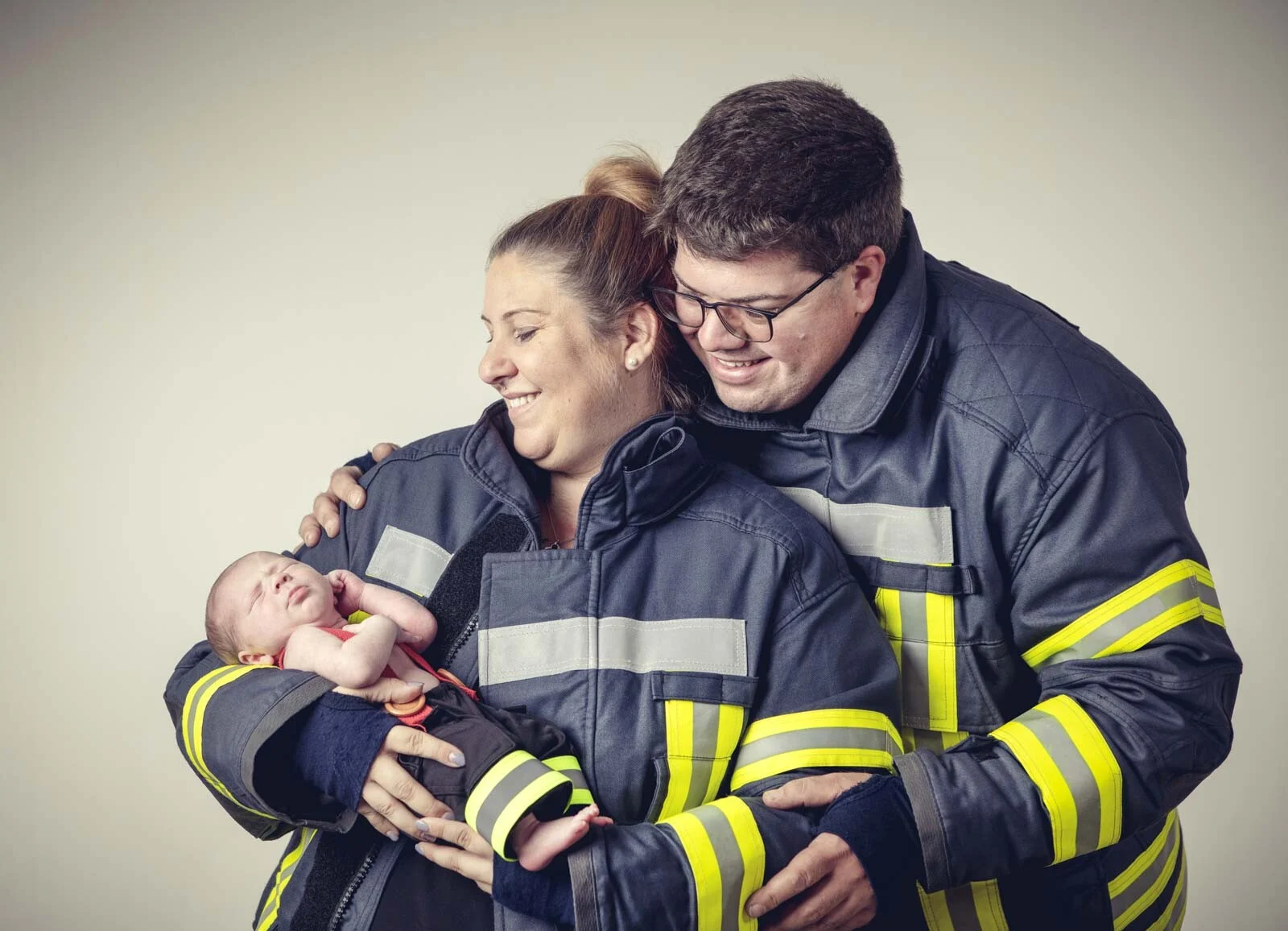 Babyfeuerwehrmann in den Armen seiner Eltern, beide in Feuerwehrjacken mit Reflexstreifen