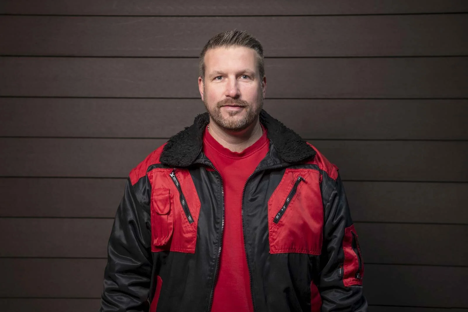 Mitarbeiter in rot-schwarzer Arbeitsjacke vor einer dunklen Holzwand