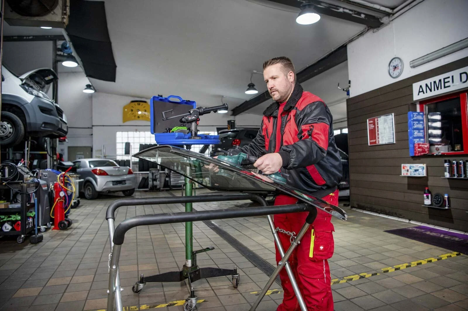 Mechaniker in roter Arbeitsjacke bearbeitet eine Autoscheibe auf einem Gestell in der Halle