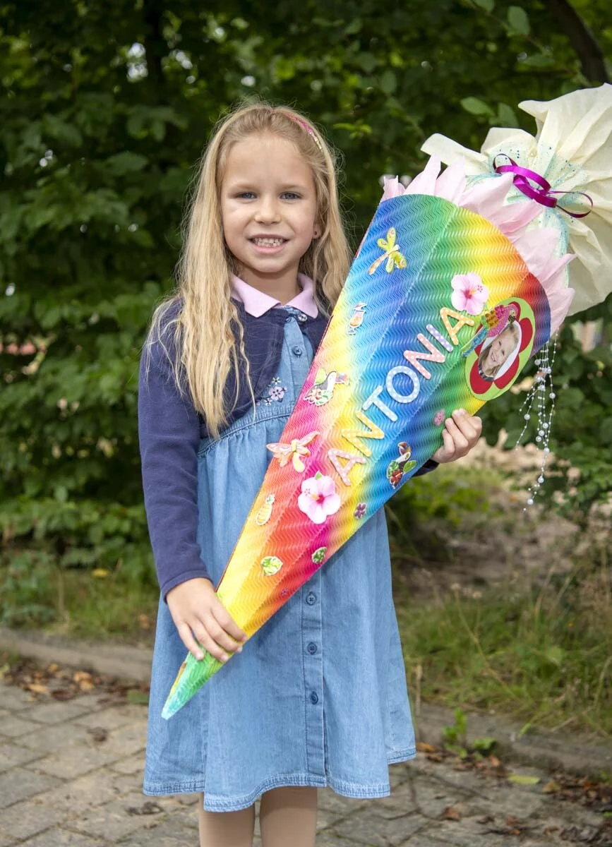 Mädchen im Jeanskleid hält eine bunte Schultüte mit Regenbogenmuster