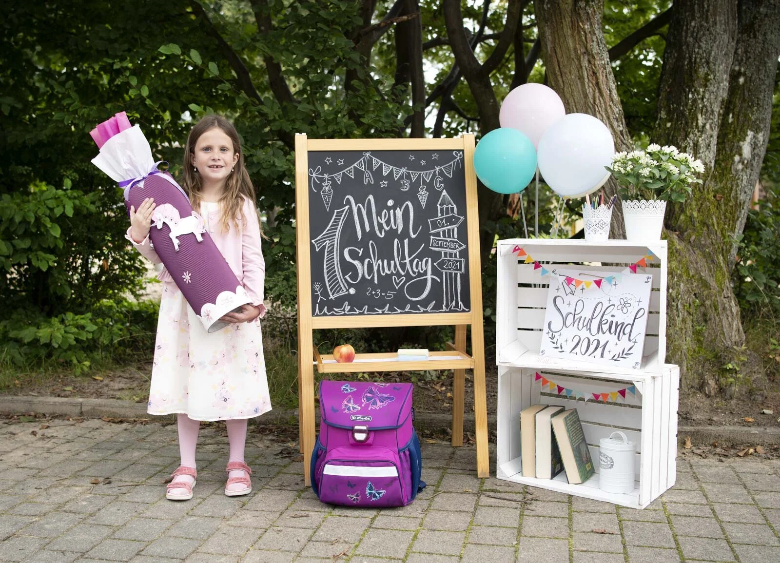 Einschulung 2021: Mädchen mit lila Schultüte vor Kreidetafel, Holzkisten und Ballons