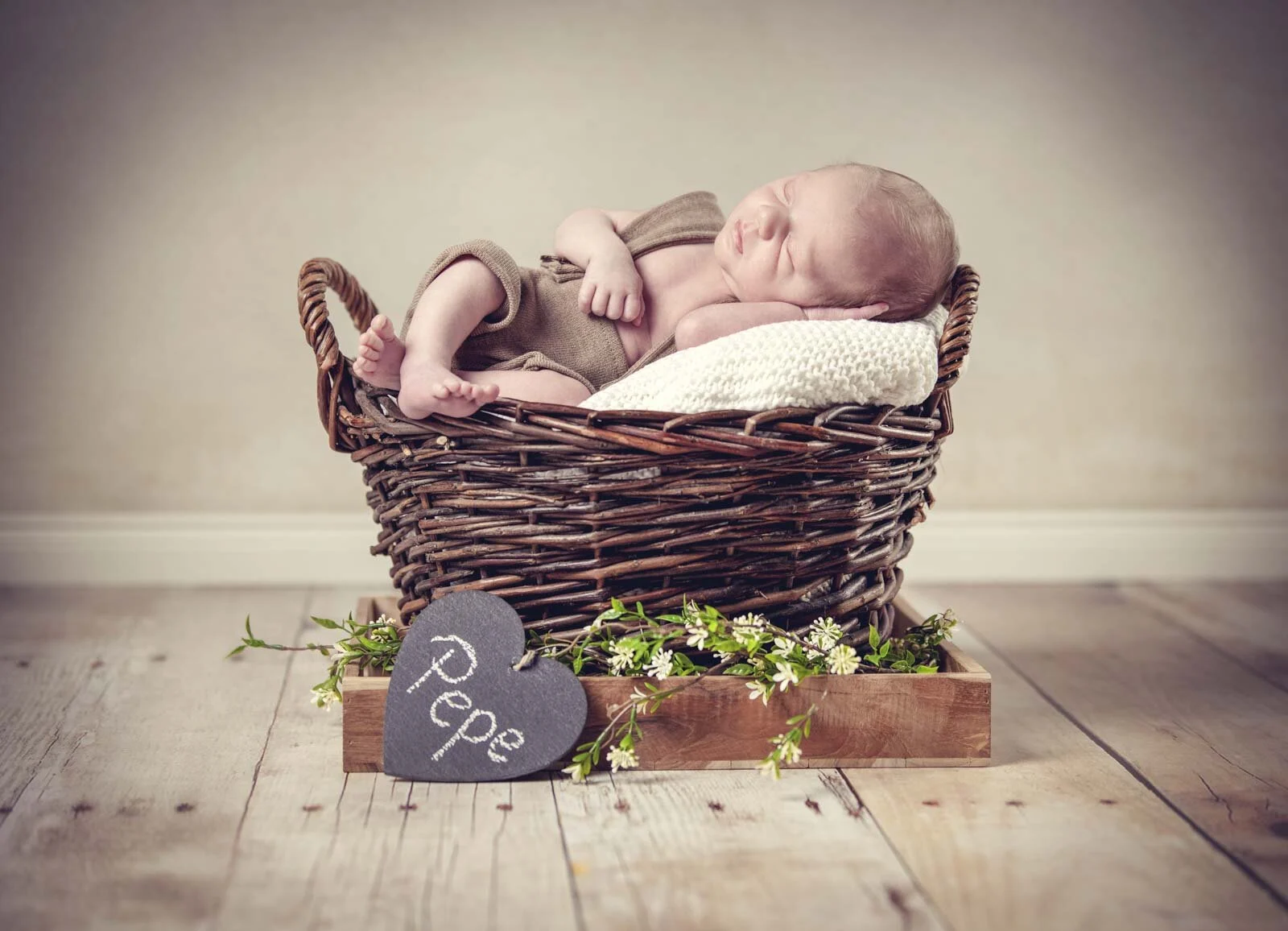 Baby schläft in einem Weidenkorb auf einer Holzkiste, davor ein Herz aus Schiefer und Blüten